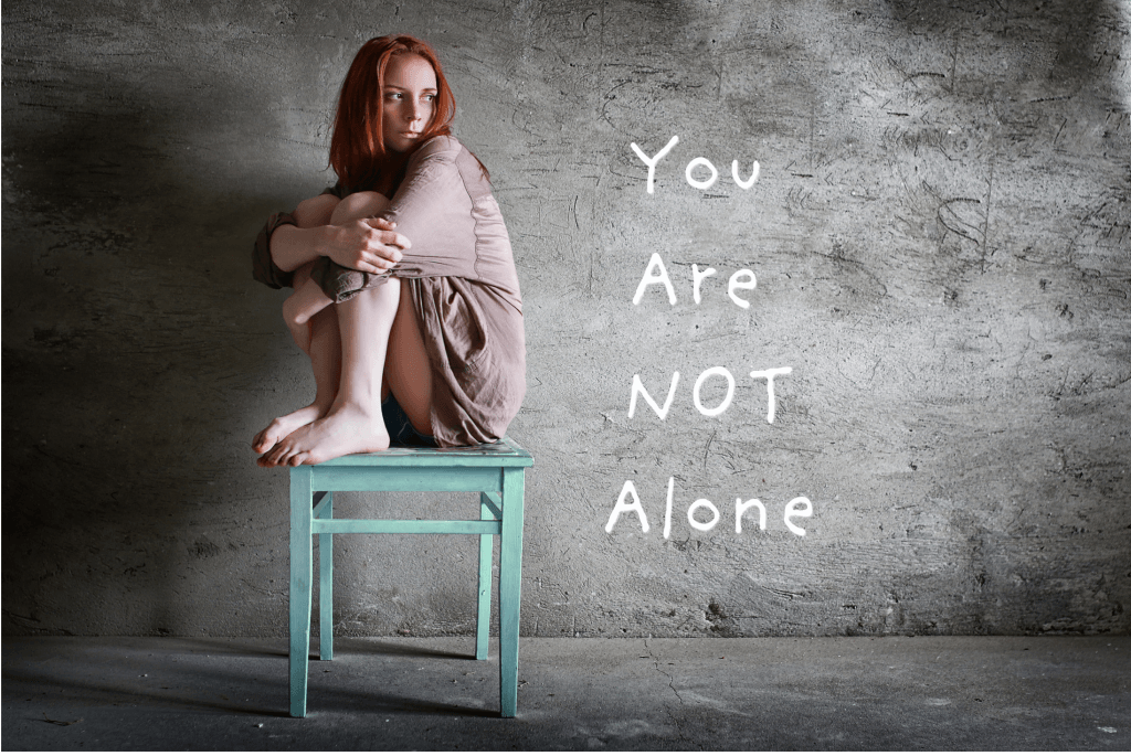 Person sitting alone with chalkboard text that reads “You Are Not Alone” – symbolizing emotional support and mental health awareness.