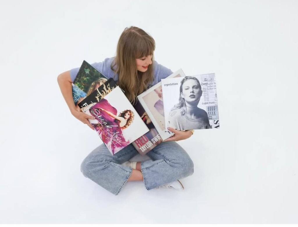 Taylor Swift Owns His Masters - aylor Swift sitting cross-legged, holding six of her studio albums with a joyful expression