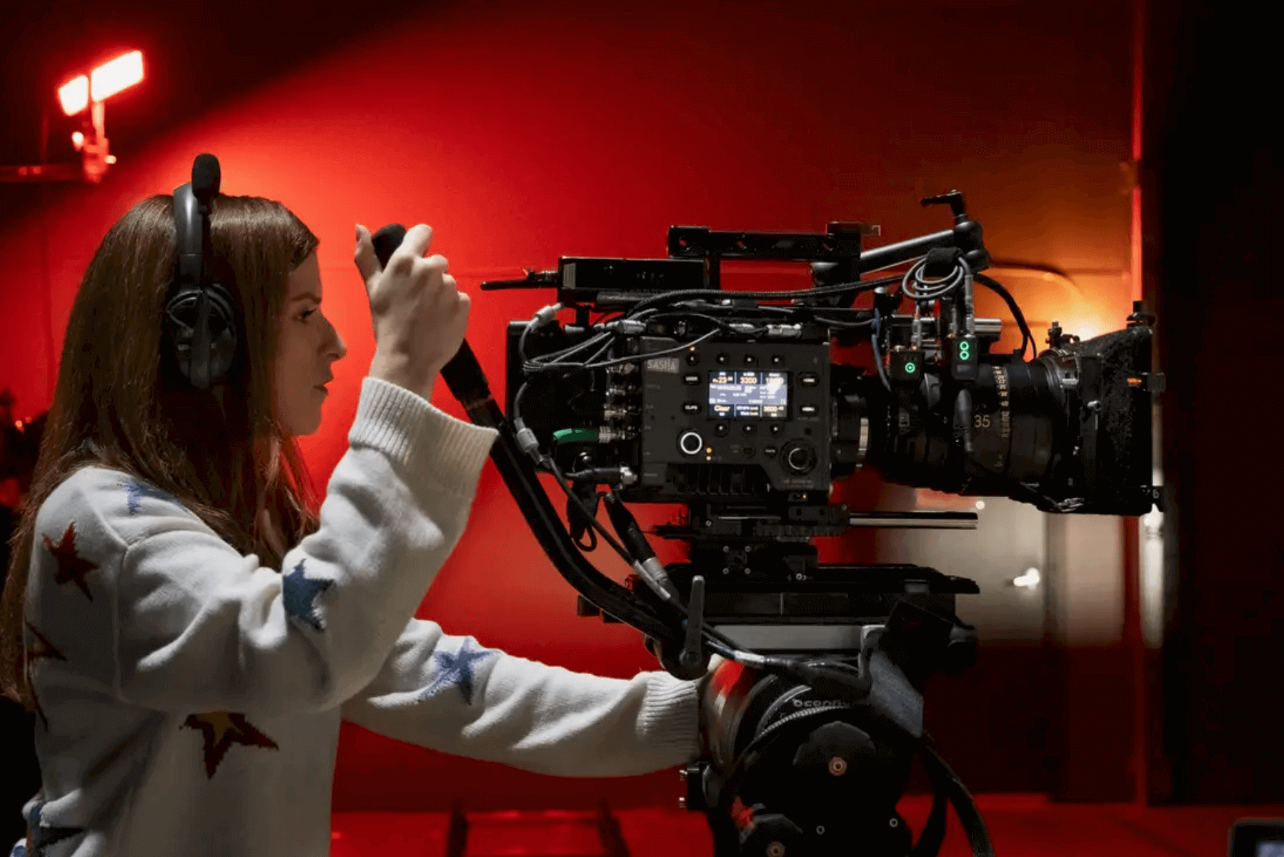 Movies directed by women - Anna Kendrick, wearing headphones and a sweater with star patterns, operates a large cinema camera on the set of her directorial debut Woman of the Hour.