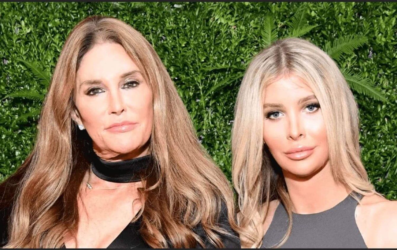 Hollywood Relationship Terms - Caitlyn Jenner and Sophia Hutchins pose together in front of a leafy green backdrop, both wearing sleek black outfits.