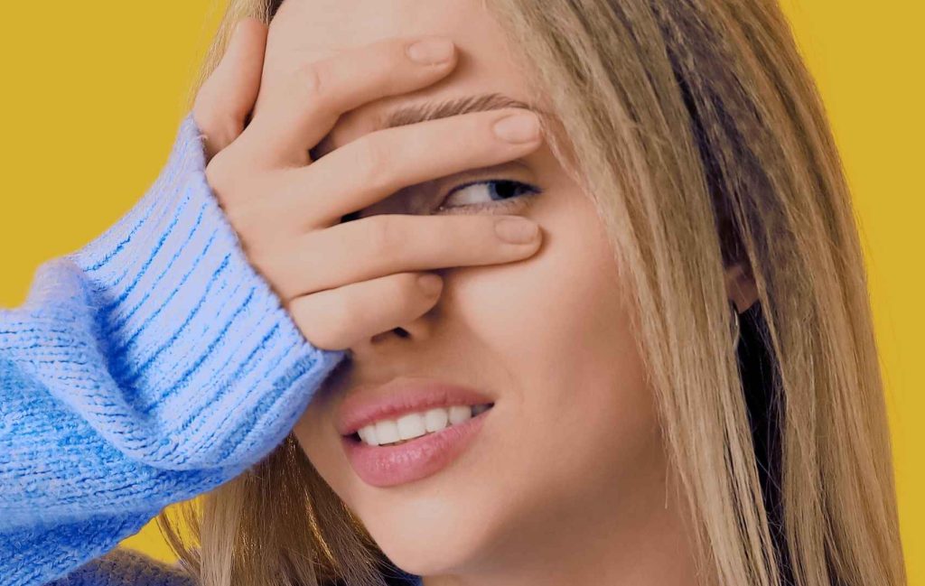 Over-Politeness - Woman half-covering her face with her hand, peeking through fingers, wearing a blue sweater on a yellow background.