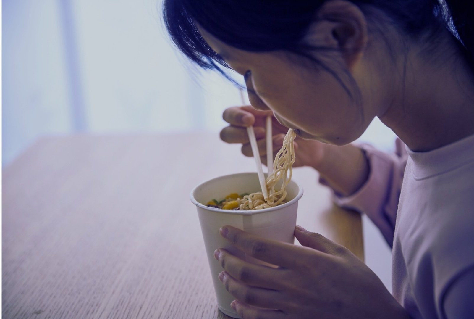 Simple life and social comparison – woman eating instant noodles from a paper cup alone at a table.